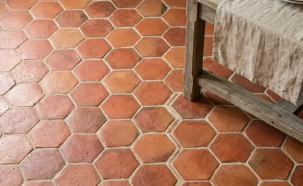 Close-up of hexagonal terracotta tomette tiles with warm red-orange patina beside a wooden table leg